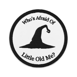 May include: Embroidered patch with a black witch's hat silhouette on a white background, encircled by a black border. The text "Who's Afraid Of Little Old Me?" is embroidered around the hat. Perfect for Halloween or adding a spooky touch to clothing or accessories.