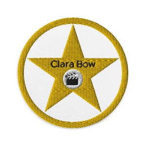 May include: Embroidered patch featuring a gold star with the text "Clara Bow" in black. The star is centered on a white circle, surrounded by a gold border. A small film clapperboard icon is at the star's center.