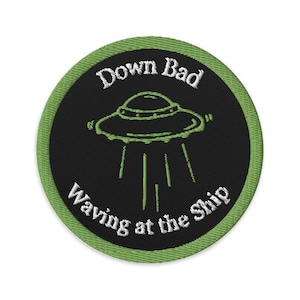 May include: Embroidered patch with a black center and a green border. The patch features a green UFO design with the text "Down Bad" above and "Waving at the Ship" below the UFO. The design is circular.