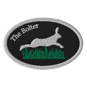 May include: Embroidered patch with an oval shape, featuring a white rabbit leaping over green grass against a black background. The text "The Bolter" is arched above the rabbit in white. The patch has a silver border.