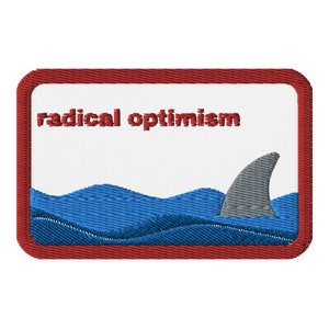 May include: Embroidered patch with a red border and the words "radical optimism" in red. The design features blue waves, a gray shark fin, and a white background.