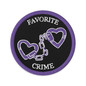 May include: Embroidered patch with a black background and a purple border. The patch features the words "FAVORITE" and "CRIME" in white, along with a design of two purple heart-shaped handcuffs connected by a chain.