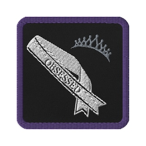 May include: Embroidered patch with a black background and a purple border. The patch features a white ribbon with the word "OBSESSED" and a small silver crown. The design is detailed and has a textured appearance.