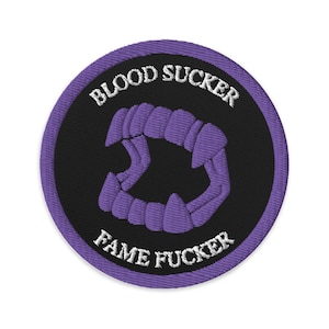 May include: Embroidered patch with a black center and a purple border. The patch features the words "BLOOD SUCKER" and "FAME FUCKER" in white, along with a purple set of fangs.