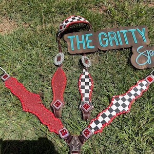 Op de afbeelding: Een set paardensportuitrusting met een rood en zwart geblokt patroon. De set bevat een hoofdstel, borsttuig en teugels, met zilveren accenten en rode edelstenen. Het bord op de achtergrond luidt "THE GRITTY Spur".