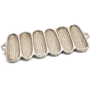 May include: A silver-colored cast iron cornbread pan with six corn-shaped molds. The pan has a rectangular shape with rounded edges and small handles on each end. The interior of each mold has a textured surface.