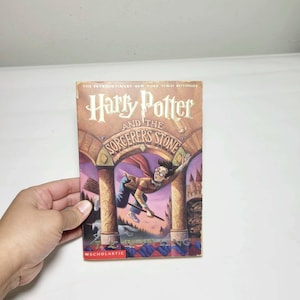 May include: A paperback copy of the book Harry Potter and the Sorcerer's Stone by J.K. Rowling. The cover features an illustration of a young character with glasses flying on a broomstick through a stone archway, with a unicorn visible below.
