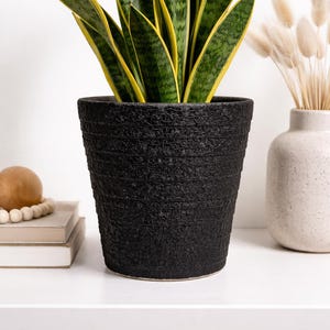 May include: A black textured ceramic plant pot holding a snake plant with green and yellow leaves. The pot is cylindrical and sits on a white surface. A decorative vase and other items are in the background.