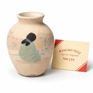 May include: A beige ceramic vase with a textured surface featuring a painted figure in black and teal. The vase has a narrow neck and a wide body. Beside the vase is a rectangular card with red diagonal stripes and the text: RANCHO FELIZ Signed Originals NM USA.