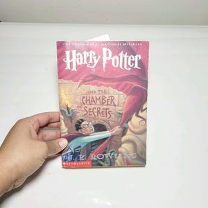 May include: A paperback book titled Harry Potter and the Chamber of Secrets by J.K. Rowling. The cover features a red phoenix, a stone wall background, and an illustration of a young person with glasses reaching toward the bird. Scholastic logo.