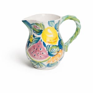 May include: White ceramic pitcher with a painted fruit design. The pitcher features a watermelon slice, lemon, and orange slices, with green and blue leaves. The handle is a twisted rope design in green. A decorative piece for serving beverages.