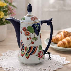 May include: A white ceramic coffee pot with a blue handle and spout, adorned with a butterfly and floral design in red, green, and orange. The pot sits on a white lace doily, with croissants and flowers in the background.