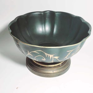 May include: A dark green decorative bowl with a scalloped rim, resting on a bronze-colored base. The bowl features gold-colored, stylized floral designs around the rim. The bowl is likely made of ceramic or porcelain.