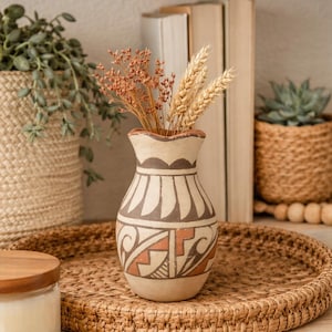 May include: A decorative ceramic vase with a Southwestern design, filled with dried flowers and wheat stalks. The vase is beige with brown and orange geometric patterns. It sits on a woven tray, with other decorative items in the background.