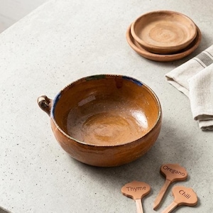 May include: A rustic, brown ceramic bowl with a handle, a blue rim, and a set of small matching plates. Three wooden plant markers labeled "Thyme", "Oregano", and "Chili" are also present, suggesting a culinary theme.