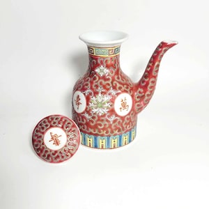 May include: Red ceramic pitcher with a spout and matching lid. The pitcher is decorated with white floral designs, Chinese characters, and a swirling pattern. The lid has a matching design. The pitcher is approximately 7 inches tall.