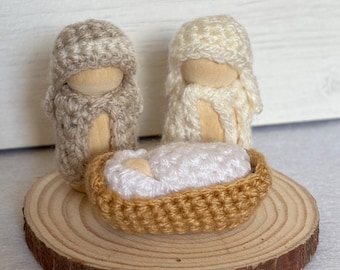 May include: A crocheted nativity scene with a brown and white figure of Mary, a white figure of Joseph, and a white baby Jesus in a brown crocheted crib. The figures are standing on a wooden base.
