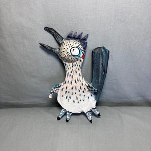 May include: A handcrafted roadrunner figurine featuring a white body with black speckles, a prominent dark blue tail, and a crest on its head. The bird has a long, pointed beak, a single visible eye with a blue outline, and small feet.
