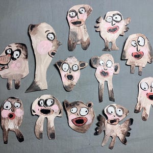 May include: A collection of eleven whimsical, hand-painted paper cutouts of expressive, cartoonish faces. Each figure has a unique design with exaggerated features, including large eyes, pink cheeks, and open mouths, in shades of brown, beige, and pink.