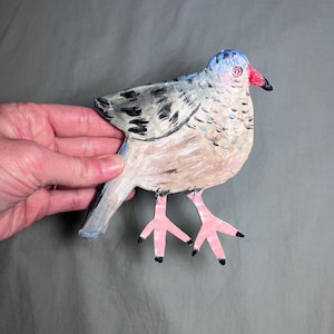 May include: A hand-painted wooden bird figurine. The bird has a blue head, a red beak, and a body with shades of tan, gray, and black. The bird's feet are pink with black tips. The figurine is held against a gray background.