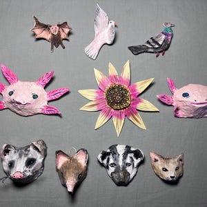 May include: A collection of handmade paper mache animal and flower ornaments. Includes a bat, dove, pigeon, axolotls, a sunflower, a possum, a raccoon, a badger, and a fox. Each ornament is painted with vibrant colors.