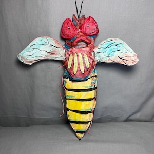 May include: A handmade, whimsical bee sculpture with a unique design. The bee features a red head with large eyes, blue and white wings, and a yellow and black striped body. The sculpture is made of paper and has a textured appearance.