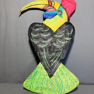 May include: A hand-carved wooden toucan sculpture. The bird features a large yellow beak, a red crest, and a body painted with black, green, and yellow. The sculpture is approximately 12 inches tall and is a decorative item.