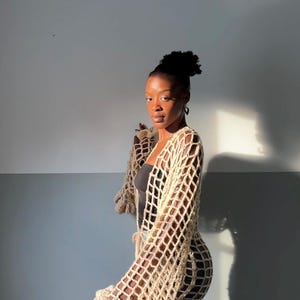 Könnte beinhalten: Ein beigefarbener Häkelcardigan mit einer langen, fließenden Silhouette und Rüschenärmeln. Der Cardigan ist aus einem leichten, offen gewebten Stoff gefertigt und hat einen Bindeverschluss in der Taille.
