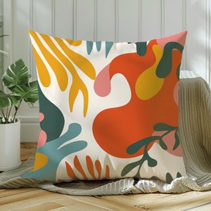 May include: A white throw pillow with a colorful abstract design featuring yellow, orange, red, pink, green, and blue shapes. The pillow is sitting on a beige throw blanket.