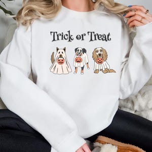 May include: White sweatshirt with the text "Trick or Treat" above three dogs dressed as ghosts, each holding a jack-o'-lantern. The dogs are different breeds, and the design is a Halloween theme.