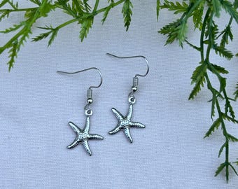 May include: A pair of silver-colored starfish earrings. Each earring features a small starfish charm suspended from a fish hook ear wire. The earrings are displayed against a white background with green foliage.