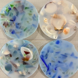 May include: Four circular coasters made of translucent resin. Two coasters feature embedded blue and green sea glass fragments, while the other two contain various small white and brown seashells, including a large central scallop shell.
