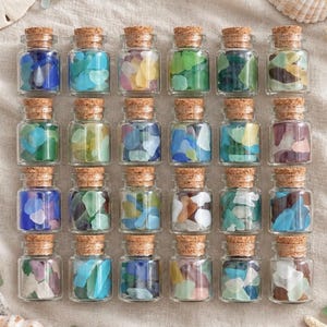 May include: A collection of twenty-four small, clear glass jars arranged in a grid, each filled with various colors of smooth sea glass and sealed with a natural cork stopper. The jars contain assorted shades of blue, green, white, and brown glass.