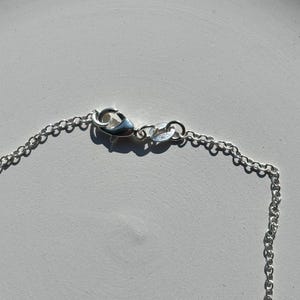 May include: Close-up of a silver necklace chain with a lobster clasp. The chain is made of small, interconnected links. The clasp is attached to a small ring with the number "925" stamped on it.