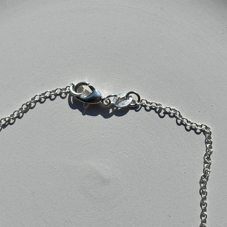 May include: Close-up of a silver necklace chain with a lobster clasp. The chain is made of small, interconnected links. The clasp is attached to a small ring with the number "925" stamped on it.