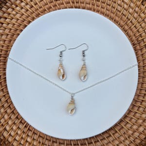 May include: A jewelry set featuring a necklace and earrings. The necklace has a small, beige seashell pendant on a silver chain. The earrings have matching seashells suspended from silver hooks. The set is displayed on a white plate against a woven background.