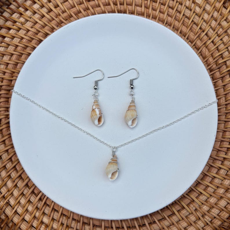 May include: A jewelry set featuring a necklace and earrings. The necklace has a small, beige seashell pendant on a silver chain. The earrings have matching seashells suspended from silver hooks. The set is displayed on a white plate against a woven background.