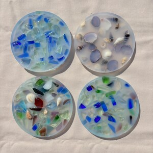 May include: Four round coasters with a clear resin base and embedded sea glass and shells. The coasters have a light blue tint and feature various colors of sea glass, including blue, green, and red. Some coasters also have small white and brown shells.