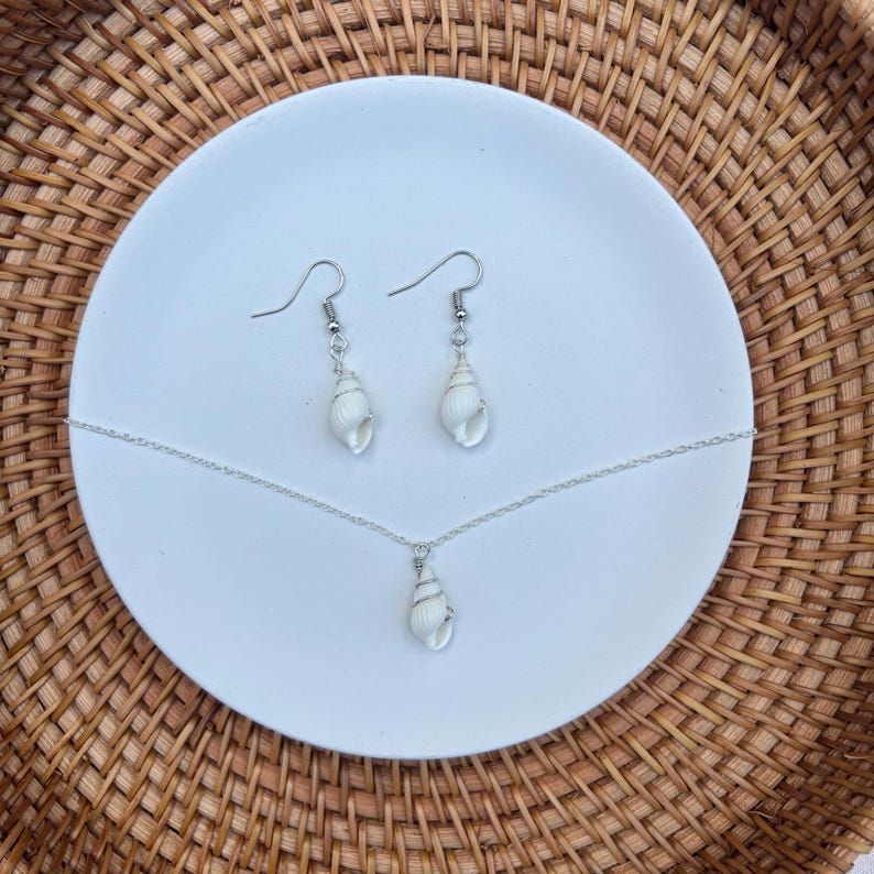 May include: A jewelry set featuring a necklace and earrings, each with a white seashell pendant. The necklace has a delicate silver chain, and the earrings have silver hooks. The set is displayed on a white plate, set against a woven background.