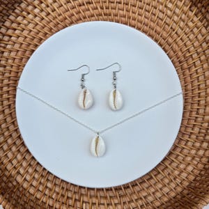 May include: A jewelry set featuring a necklace and earrings with natural white seashell pendants. The earrings are drop style, and the necklace has a single pendant. The set is displayed on a white plate against a woven background.