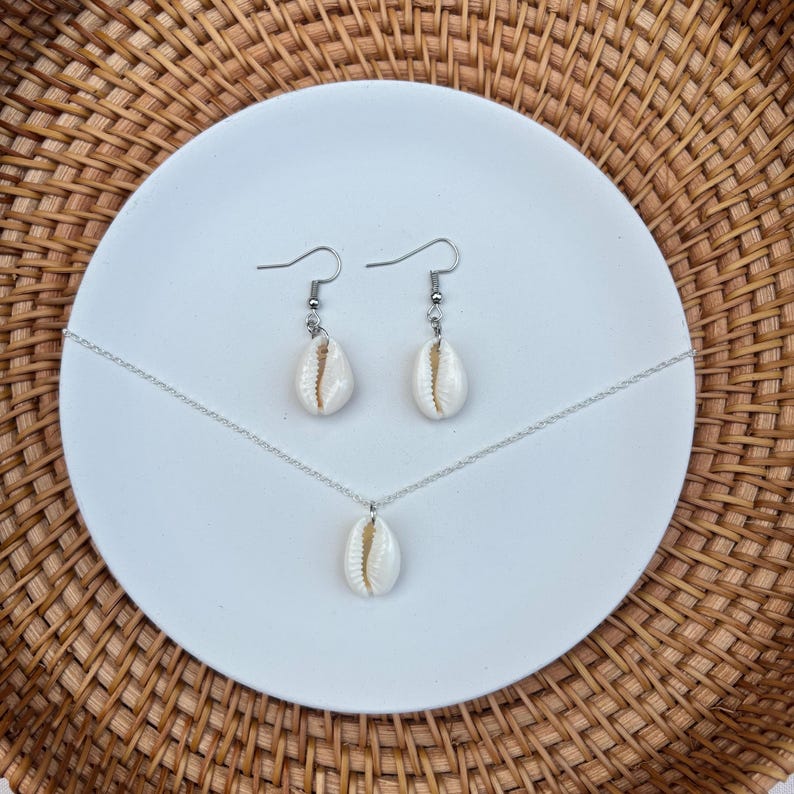 May include: A jewelry set featuring a necklace and earrings with natural white seashell pendants. The earrings are drop style, and the necklace has a single pendant. The set is displayed on a white plate against a woven background.