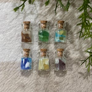 May include: Six small glass bottles with cork stoppers, each filled with colorful, irregularly shaped pieces. The bottles contain shades of brown, green, blue, yellow, and purple. The bottles are arranged on a soft, textured surface.