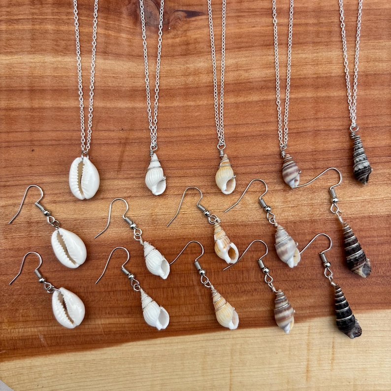 May include: A collection of seashell jewelry, including necklaces and earrings. The necklaces feature various seashells suspended from delicate silver chains. The earrings have matching seashell designs. The shells vary in shape, size, and color, with some displaying natural patterns.