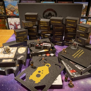 Puede incluir: Un organizador de juegos de mesa con varios componentes, incluyendo figuras en miniatura, dados y tableros de juego. El conjunto incluye cajas negras etiquetadas con términos relacionados con el juego como "Sun Stalker" y "Fighting Arts".