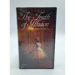 Könnte beinhalten: Versiegelte DVD-Hülle für "The Truth of Illusion" von Cirque du Soleil. Das Cover zeigt eine Frau in einem cremefarbenen Kleid und einem goldfarbenen Käfigrock, vor einem warmen, verzierten Hintergrund. Der Titel ist in eleganter Schrift gehalten.