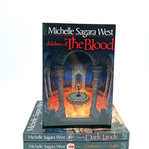 May include: A stack of three books by Michelle Sagara West. The top book is titled "children of The Blood" with a dark cover illustration of a figure in a fantasy setting. The other two books are titled "into the Dark Lands" and "chains of Darkness, chains of Light".