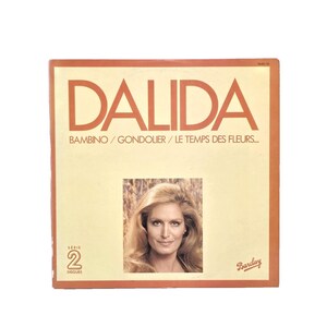 May include: A vintage vinyl record album cover featuring the name "DALIDA" in large orange letters. Below the name are the song titles "BAMBINO / GONDOLIER / LE TEMPS DES FLEURS...". A portrait of a woman with blonde hair is in the center. The album is a "SERIE 2 DISQUES" release.