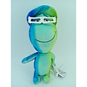 May include: A plush toy character from the film Soul, in shades of blue and green. The character has white-framed glasses and a matching hat, with a smiling face. A tag is attached to the toy.