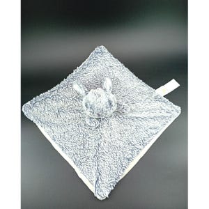 Peut inclure: Une couverture de sécurité pour bébé en peluche grise et blanche avec une petite tête de lapin au centre. La couverture est de forme carrée avec une surface douce et texturée et une bordure blanche. Le lapin a de longues oreilles et un petit visage rond.