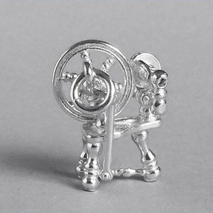 May include: A silver spinning wheel charm with a detailed design. The wheel is turned by a small handle and has a spindle for winding yarn.